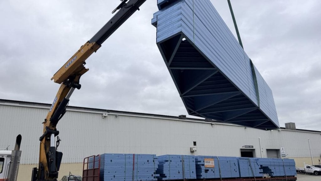 Flat-packed light gauge steel wall frames and roof trusses being craned onto an iBuild delivery truck in labelled bundles